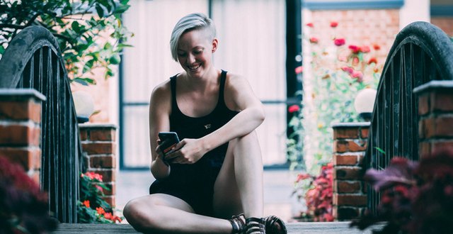 Alida Svensson, en ung kvinna sitter på en bro och tittar på en mobiltelefon som hon har i händerna.