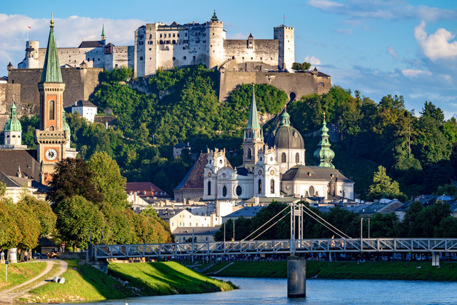 Vy från Salzburg med en stor borg på ett berg i bakgrunden, en flod i förgrunden, kyrkor och byggnader med tinnar och torn, samt bostadshus vid floden.