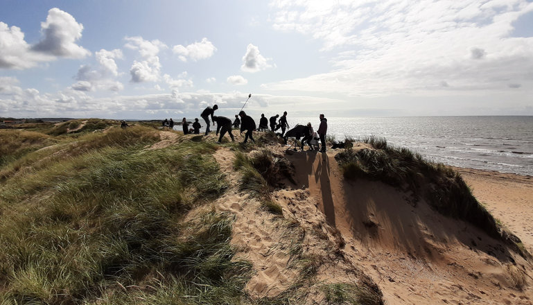 Filminspelning med flera personer på toppen av en sanddyn vid havet.
