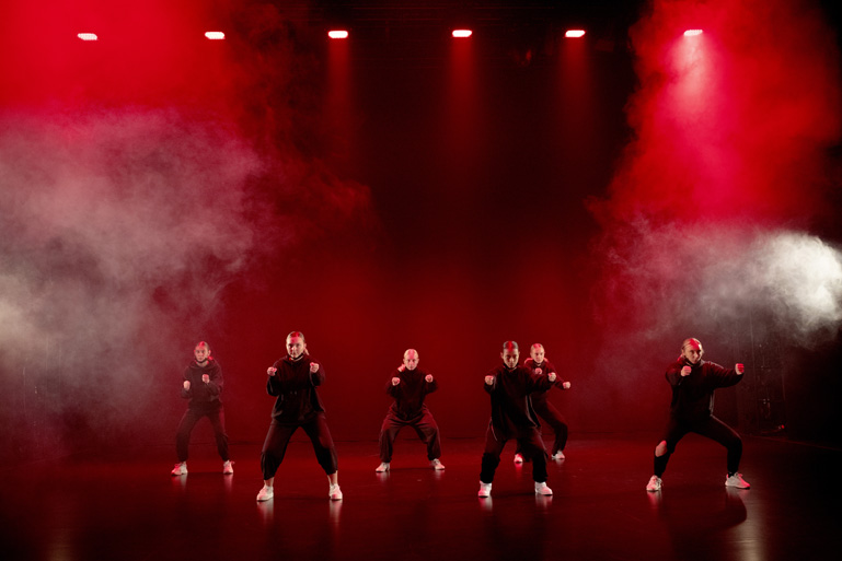 Dansgrupp i svarta kläder på en scen med rök och upplyst med rött ljus.
