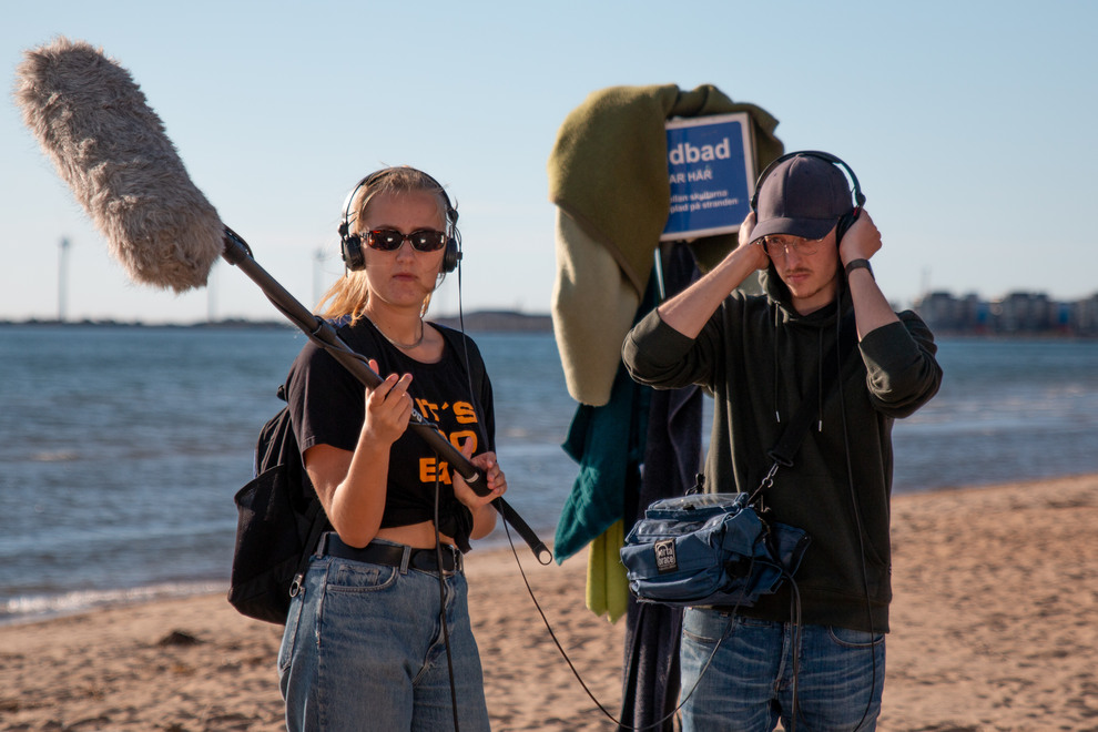 Två personer med utrustning för filminspelning på stranden.