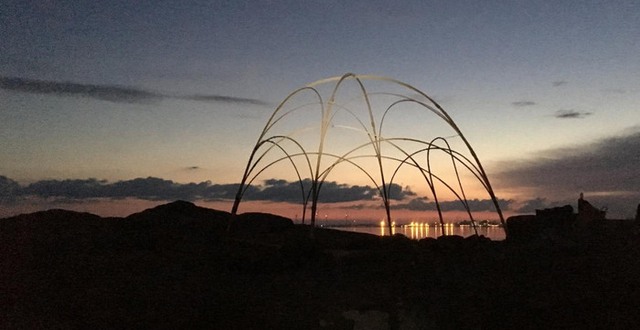 Ett konstverk skapat av sammanfogade träbågar syns som en siluett mot havet och kvällshimlen.