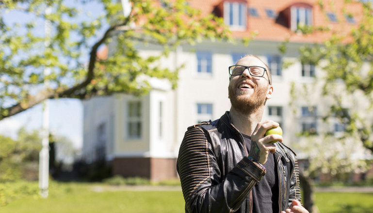 En kille med äpple i handen står i en park framför vit skolbyggnad.