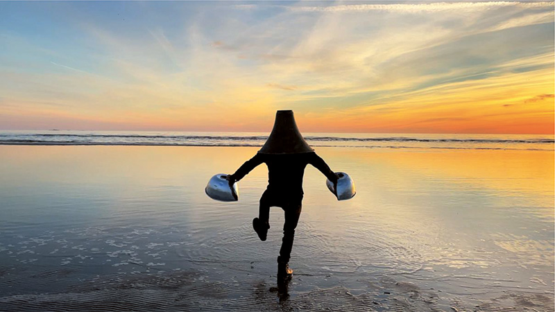 En person med en strut över huvudet och sivriga klumpar på händerna kliver med stora steg ut i havet i en stilla solnedgång.