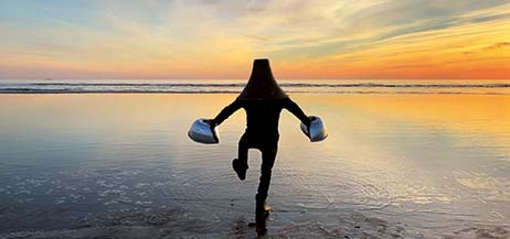 En person med en strut över huvudet och sivriga klumpar på händerna kliver med stora steg ut i havet i en stilla solnedgång.
