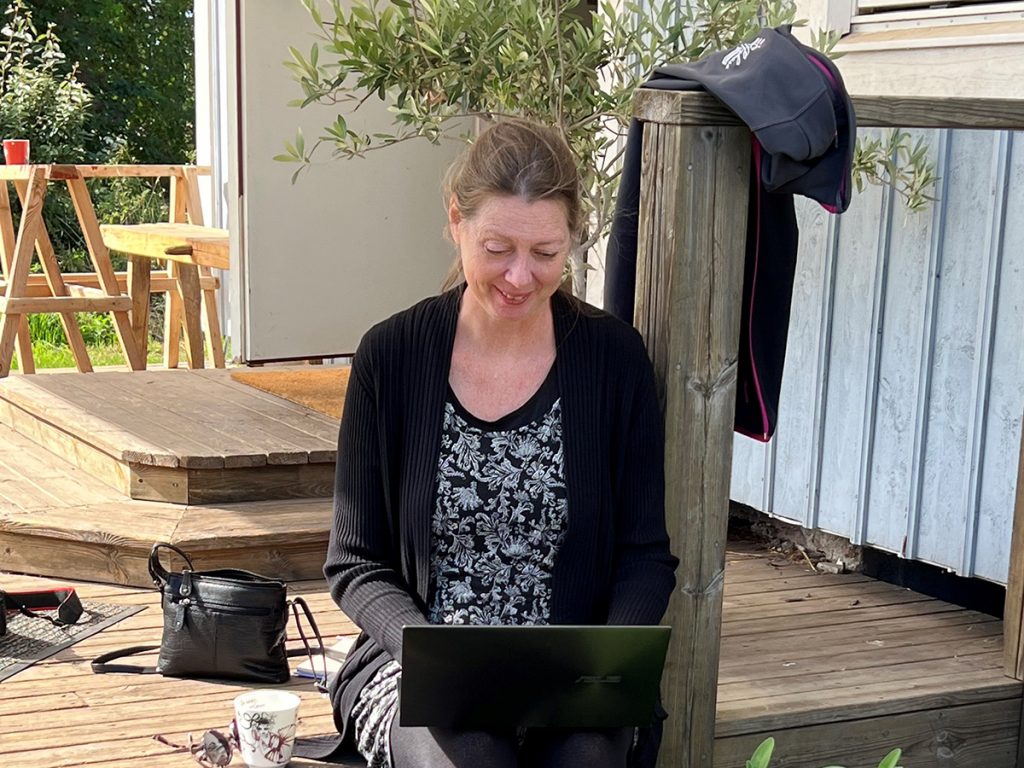 En kvinna sitter på en veranda och skriver med en laptop i knät.