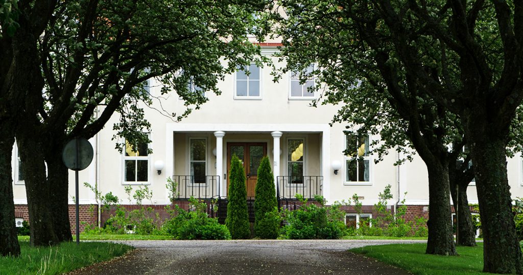 Den gräddvita huvudbyggnaden på Katrinebergs folkhögskola sedd från allén som leder fram till entrén.