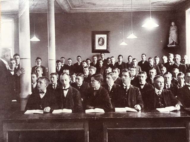 Svartvit äldre bild med unga män i en lektionssal.