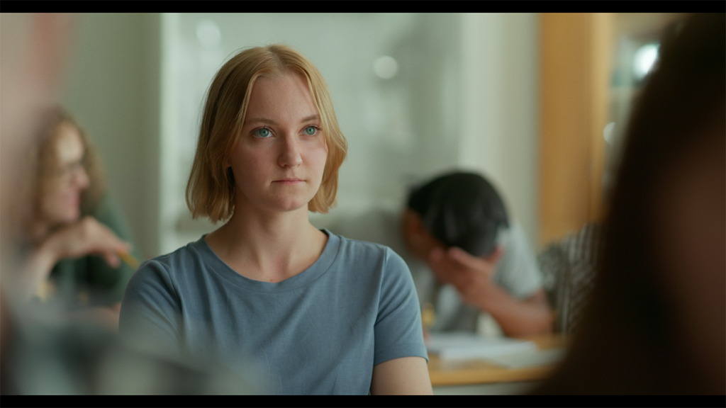 Klipp ur filmen Ikaros, en tjej som tittar mot kameran men med blicken fäst ovanför betraktaren, i bakgrunden sitter två personer i skolbänkar.