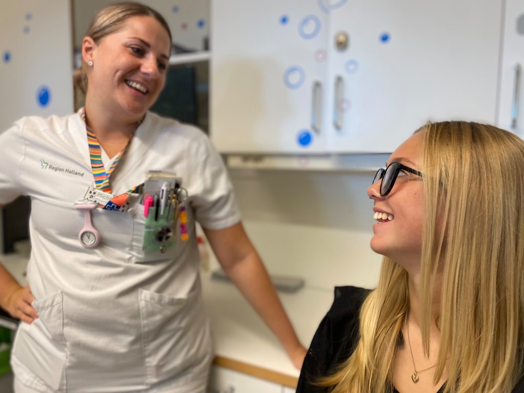 Sjuksköterska Michaela Ulfsparre och Lova Herrlin i provtagningsrum. Tittar leende på varandra.