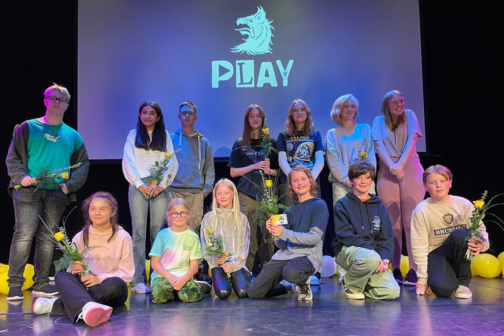 Gruppfoto med barn och ungdomar som håller i blombuketter, de står uppställda framför en filmduk med texten "PLAY".
