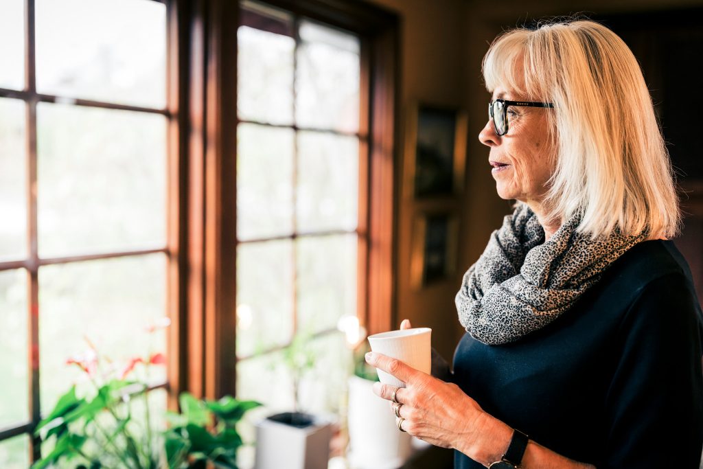 Äldre kvinna tittar ut genom fönstret och håller en kaffekopp i handen