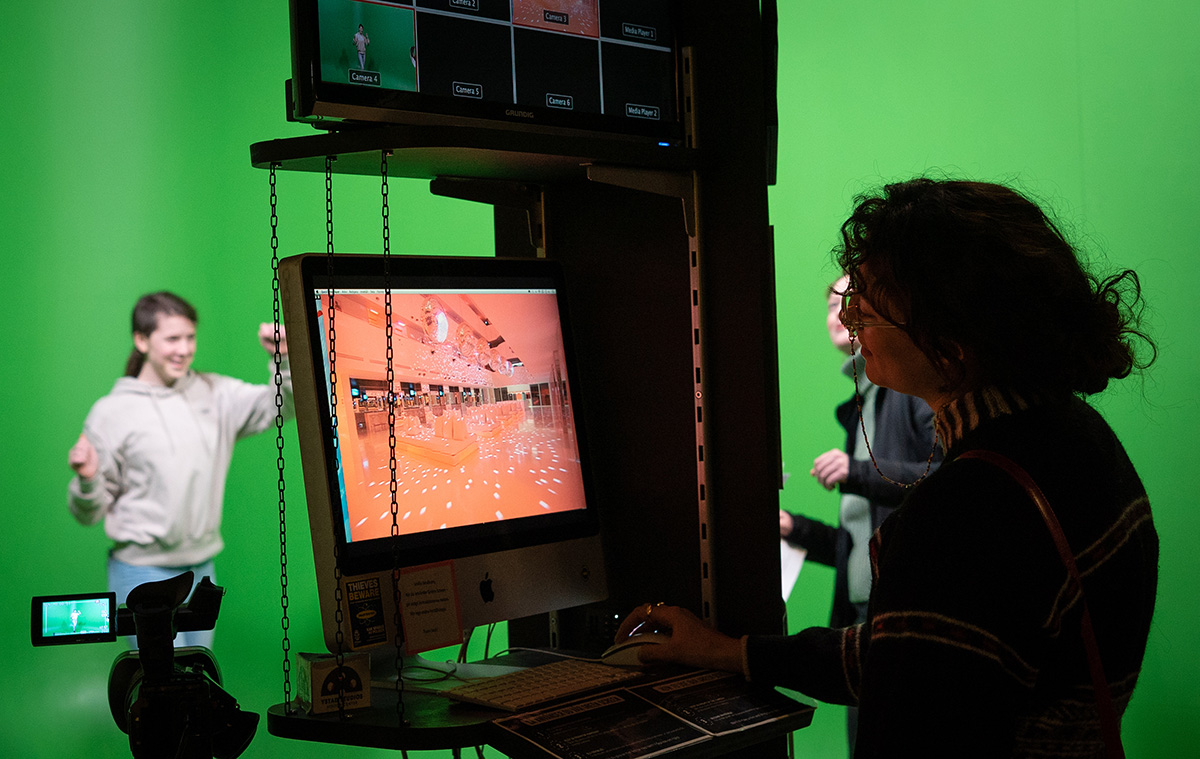 Filminspelning med greenscreen där ett barn agerar framför en grön vägg och en vuxen står och tittar på en datorskärm som visar en annan bakgrundsmiljö.