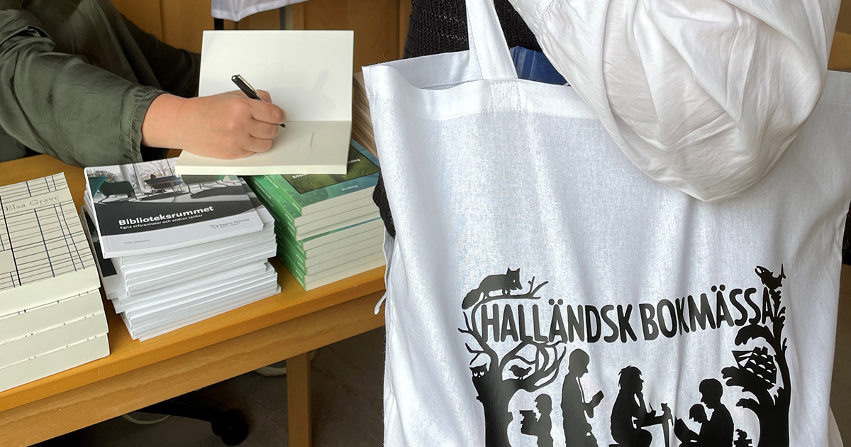 Närbild på en hand som skriver på försättsbladet i en bok och en annan person som bär en tygkasse med texten "Halländsk bokmässa" som står framför.