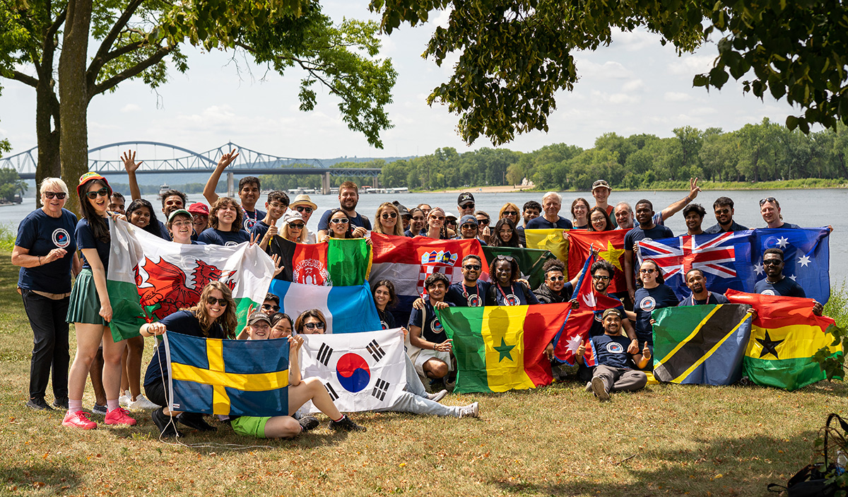 Gruppfotografi med cirka 50 unga personer som står vid en flod och håller i flaggor från flera olika länder.