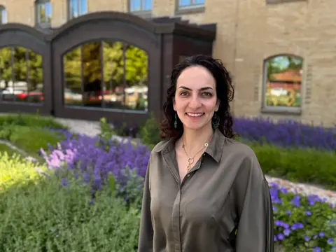 Leende kvinna med mörkt hår och brun blus står framför tegelhus. Blommor i bakgrunden.